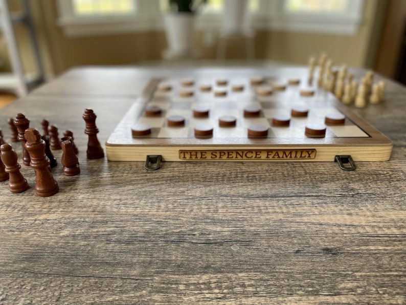 K&ouml;nnte beinhalten: Ein Schach- und Dame-Kombinationsspiel aus Holz mit dunkelbraunen und hellen Holzfiguren. Auf dem Spiel steht der Text "THE SPENCE FAMILY". Das Spiel steht auf einem Holztisch.