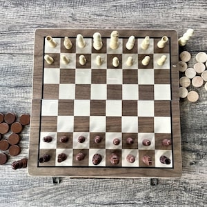 K&ouml;nnte beinhalten: Ein zusammenklappbares Schach- und Dame-Set aus Holz. Das Schachbrett ist braun und wei&szlig;, mit Schachfiguren aus hellem und dunklem Holz. Dame-Spielsteine sind braun und helles Holz. Das Set ist ge&ouml;ffnet und spielbereit.
