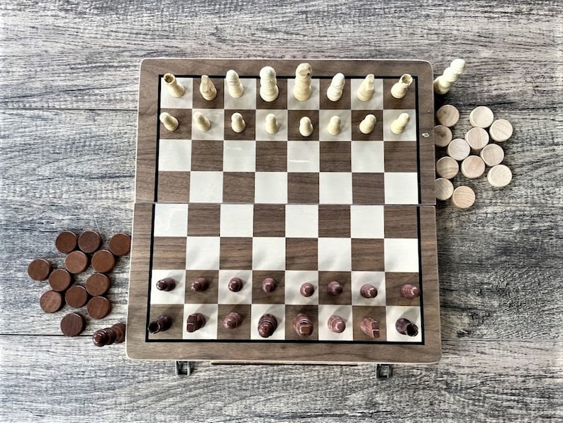 K&ouml;nnte beinhalten: Ein zusammenklappbares Schach- und Dame-Set aus Holz. Das Schachbrett ist braun und wei&szlig;, mit Schachfiguren aus hellem und dunklem Holz. Dame-Spielsteine sind braun und helles Holz. Das Set ist ge&ouml;ffnet und spielbereit.