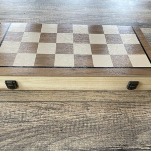 K&ouml;nnte beinhalten: Ein Schachspiel aus Holz mit einem hellbraunen und cremefarbenen Schachbrett. Das Set befindet sich in einer geschlossenen, rechteckigen Box mit dunklem Rand und Metallverschl&uuml;ssen. Die Holzmaserung ist sichtbar.