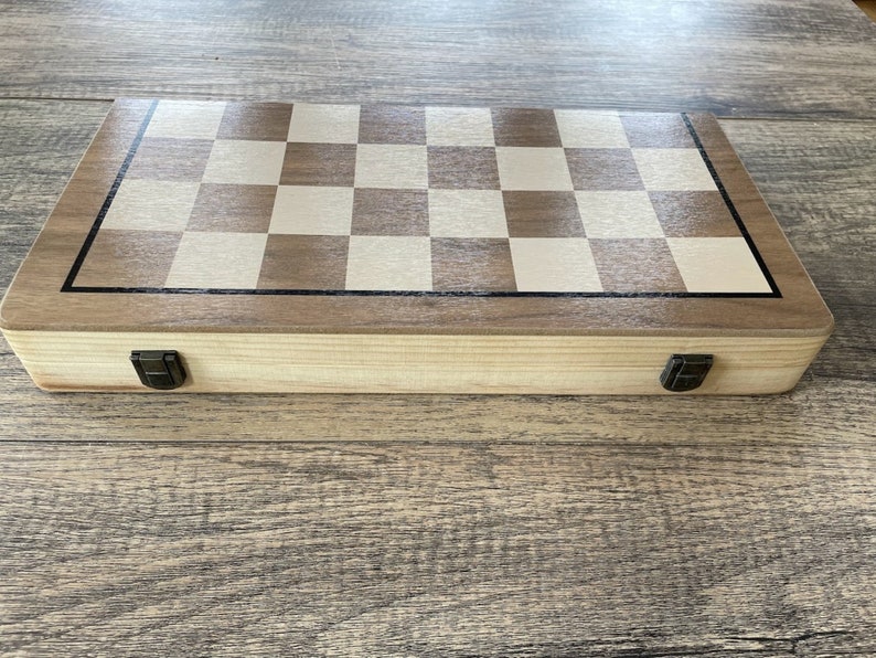 K&ouml;nnte beinhalten: Ein Schachspiel aus Holz mit einem hellbraunen und cremefarbenen Schachbrett. Das Set befindet sich in einer geschlossenen, rechteckigen Box mit dunklem Rand und Metallverschl&uuml;ssen. Die Holzmaserung ist sichtbar.