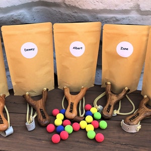 May include: Five personalized wooden slingshots with names engraved on them,  along with a bag of colorful balls. The slingshots are brown and have a white elastic band. The balls are red, blue, green, and yellow.
