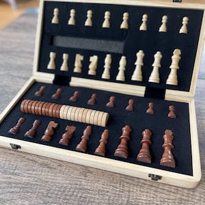 K&ouml;nnte beinhalten: Ein Schachspiel aus Holz mit einem hellfarbigen Holzrahmen und einem schwarzen Filzinnenraum. Die Schachfiguren sind in zwei Farben gehalten: hellbeige und dunkelbraun. Das Set ist ge&ouml;ffnet und zeigt die Anordnung der Figuren.