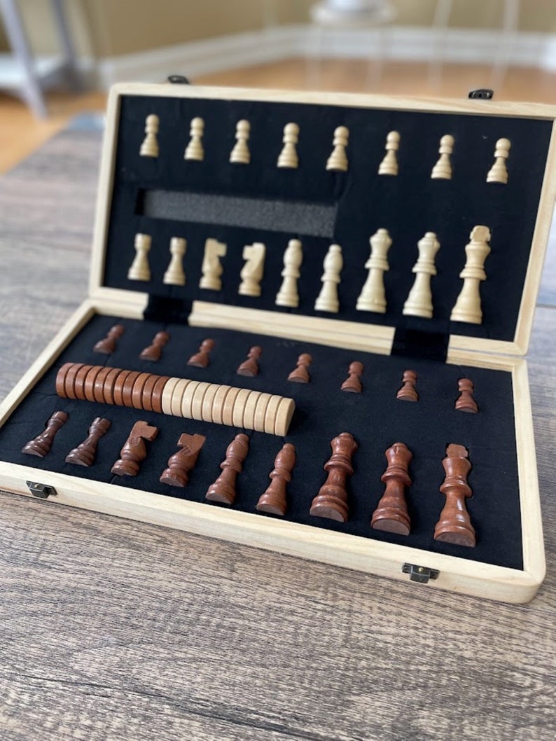 K&ouml;nnte beinhalten: Ein Schachspiel aus Holz mit einem hellfarbigen Holzrahmen und einem schwarzen Filzinnenraum. Die Schachfiguren sind in zwei Farben gehalten: hellbeige und dunkelbraun. Das Set ist ge&ouml;ffnet und zeigt die Anordnung der Figuren.