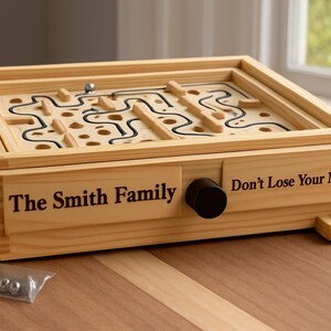 Puede incluir: Juego de laberinto de madera con dise&ntilde;o de laberinto y pomo negro. El texto "The Smith Family" y "Don't Lose Your Marbles" est&aacute;n impresos en la parte delantera. Incluye una peque&ntilde;a bolsa de canicas met&aacute;licas. El juego est&aacute; sobre una superficie de madera.