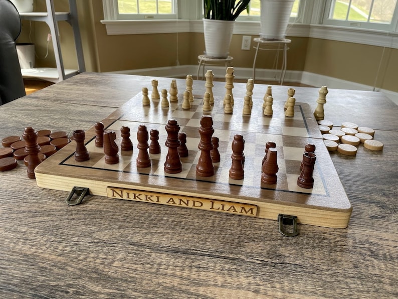 K&ouml;nnte beinhalten: Ein Schach- und Dame-Set aus Holz mit braunen und beigen Figuren. Das Schachbrett hat einen hellen Holzrahmen und der Text "NIKKI AND LIAM" ist auf der Vorderseite eingraviert. Das Set steht auf einem Holztisch.