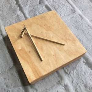 May include: A wooden square board with two wooden sticks and a small rake on top. The board is light brown and has a smooth surface.