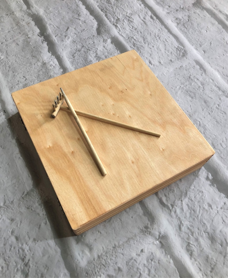 May include: A wooden square board with two wooden sticks and a small rake on top. The board is light brown and has a smooth surface.