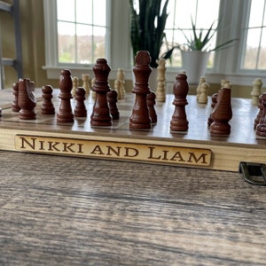 K&ouml;nnte beinhalten: Ein Schachspiel aus Holz mit braunen und beigen Figuren auf einem Schachbrett. Das Brett befindet sich in einem Holzkoffer mit Metallverschl&uuml;ssen. Der Name "Nikki and Liam" ist auf einer Holzplakette eingraviert.