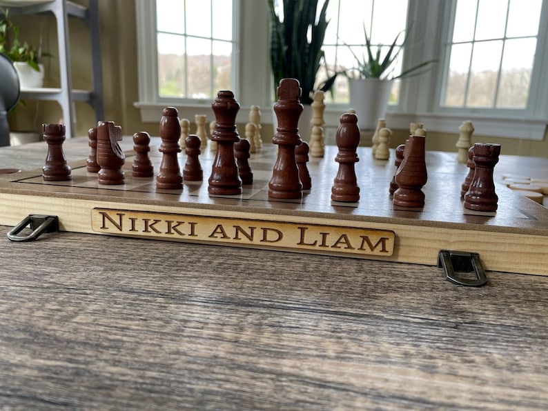 K&ouml;nnte beinhalten: Ein Schachspiel aus Holz mit braunen und beigen Figuren auf einem Schachbrett. Das Brett befindet sich in einem Holzkoffer mit Metallverschl&uuml;ssen. Der Name "Nikki and Liam" ist auf einer Holzplakette eingraviert.