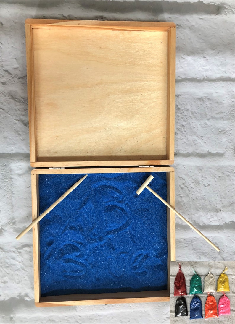 May include: A wooden box with a blue sand tray inside. The box is open and the sand tray is visible. There are two wooden tools for drawing in the sand. There are eight small bags of colored sand next to the box.