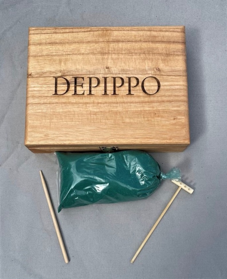 May include: A wooden box with the name "DEPIPPO" engraved on the lid. The box contains a bag of green sand, two wooden sticks, and a small wooden rake.