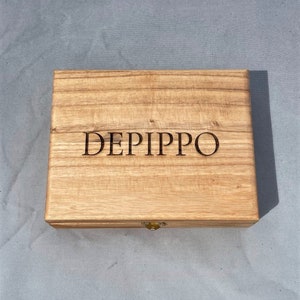May include: A wooden box with a hinged lid and a brass latch. The box is made of light wood with a natural grain. The name "DEPIPPO" is engraved on the top of the box.