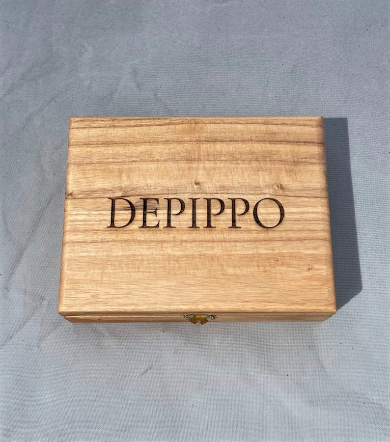 May include: A wooden box with a hinged lid and a brass latch. The box is made of light wood with a natural grain. The name "DEPIPPO" is engraved on the top of the box.