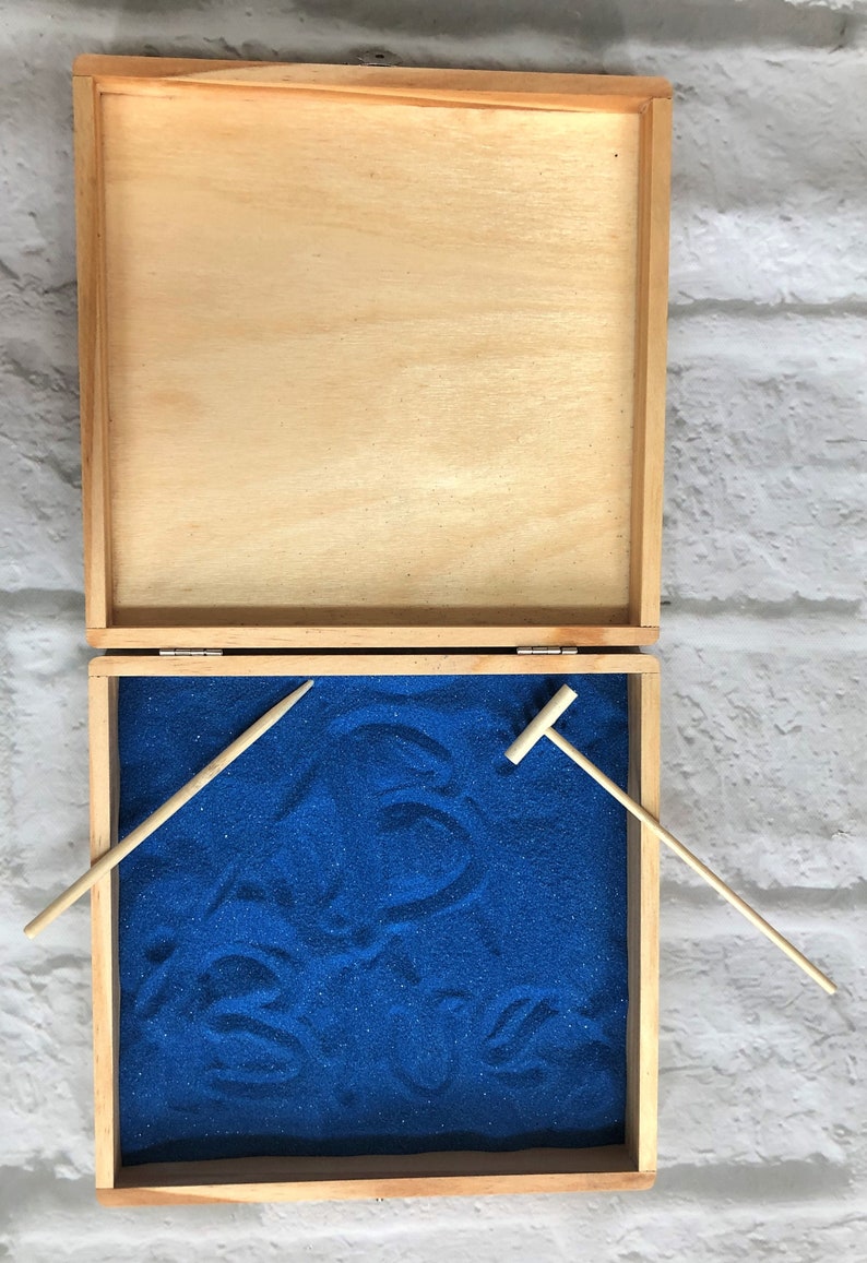 May include: A wooden box with a blue sand tray and two wooden tools. The box is open and the sand tray is visible. The tools are a rake and a small shovel.
