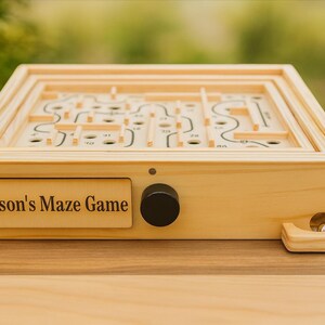 Puede incluir: Un juego de laberinto de madera con el texto "Jaxson's Maze Game" en la parte delantera. El juego presenta una perilla negra y una peque&ntilde;a bola de metal. El laberinto es una serie de caminos y agujeros.