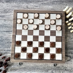 K&ouml;nnte beinhalten: Ein h&ouml;lzernes Damebrett mit einem braun-wei&szlig;en Schachmuster. Das Brett ist ge&ouml;ffnet, mit braunen und hellfarbigen Dame-Spielsteinen. Zus&auml;tzliche Spielsteine ​​sind um das Brett verstreut, und Schachfiguren befinden sich in der oberen rechten Ecke.