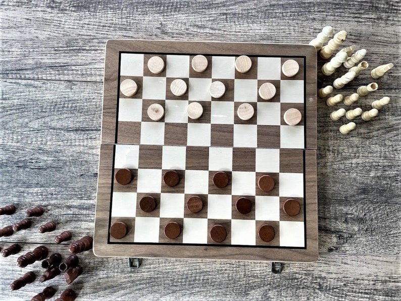 K&ouml;nnte beinhalten: Ein h&ouml;lzernes Damebrett mit einem braun-wei&szlig;en Schachmuster. Das Brett ist ge&ouml;ffnet, mit braunen und hellfarbigen Dame-Spielsteinen. Zus&auml;tzliche Spielsteine ​​sind um das Brett verstreut, und Schachfiguren befinden sich in der oberen rechten Ecke.