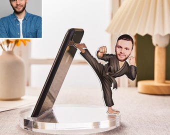 May include: A clear acrylic phone stand with a cartoon cutout of a person in a karate pose. The phone is black and angled against the stand. The cutout is wearing a black karate uniform.