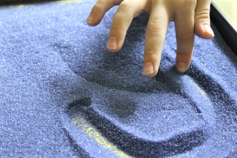 May include: A hand is making a design in a tray of blue sand.