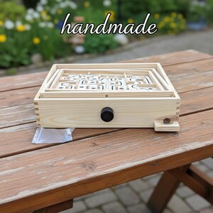Puede incluir: Un juego de laberinto de madera hecho a mano con una perilla de control negra. El juego est&aacute; sobre una mesa de madera al aire libre, con la palabra "handmade" escrita en la parte superior. El juego presenta un dise&ntilde;o de laberinto.