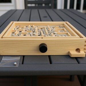 Puede incluir: Un juego de laberinto de madera con una perilla negra y una peque&ntilde;a bola de metal en una bolsa de pl&aacute;stico transparente. El juego presenta un dise&ntilde;o de laberinto con l&iacute;neas y agujeros negros. El marco de madera es de color claro.