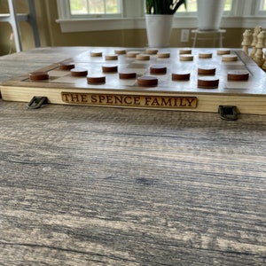 K&ouml;nnte beinhalten: Schach- und Damebrett aus Holz mit dem eingravierten Text "THE SPENCE FAMILY" an der Seite. Das Brett hat braune und nat&uuml;rliche Holz-Dame- und Schachfiguren. Das Brett liegt auf einem Holztisch.