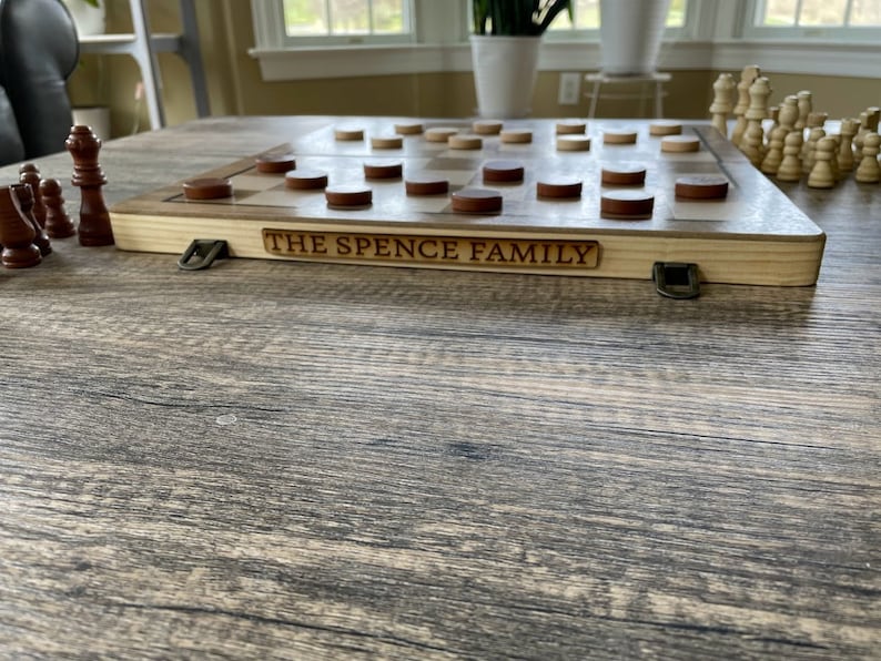 K&ouml;nnte beinhalten: Schach- und Damebrett aus Holz mit dem eingravierten Text "THE SPENCE FAMILY" an der Seite. Das Brett hat braune und nat&uuml;rliche Holz-Dame- und Schachfiguren. Das Brett liegt auf einem Holztisch.