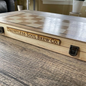 K&ouml;nnte beinhalten: Ein Schach- und Backgammon-Set aus Holz mit hellem Holzfinish. Das Set hat ein Schachbrettmuster und den Text "Dauntless Soul Brew Co." auf einer Holzplakette. Ein Metallverschluss sichert das Set.