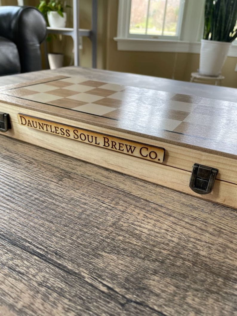 K&ouml;nnte beinhalten: Ein Schach- und Backgammon-Set aus Holz mit hellem Holzfinish. Das Set hat ein Schachbrettmuster und den Text "Dauntless Soul Brew Co." auf einer Holzplakette. Ein Metallverschluss sichert das Set.