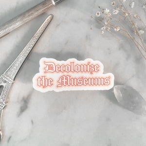 May include: A white sticker with the text "Decolonize the Museums" in red and orange lettering. The sticker is on a marble surface.