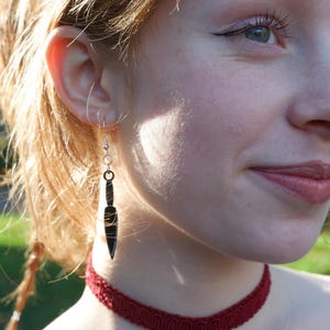 May include: A woman wearing a red choker necklace with a heart pendant. She is wearing a pair of silver earrings with a knife-shaped charm.