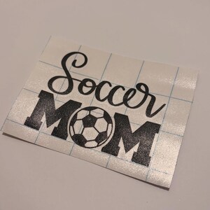 May include: A black vinyl decal on a white grid transfer paper featuring the word Soccer in a cursive script font above the word MOM in a bold block font, with a soccer ball graphic replacing the letter O in the word MOM.