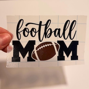 May include: A vinyl decal featuring the black script text football above the block letter M, a brown football graphic replacing the letter O, and the block letter M. The design is printed on a white backing sheet with a blue grid pattern.