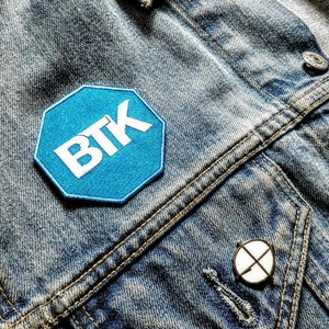 May include: Close-up of a denim jacket with a blue and white "BTK" patch, a pin, and a green "SUMMERDALE AV" pin. The jacket has a classic design with visible stitching and metal buttons.
