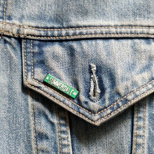 May include: A green enamel pin with white lettering that reads "SUMMERDALE AV" is pinned to a blue denim jacket pocket.