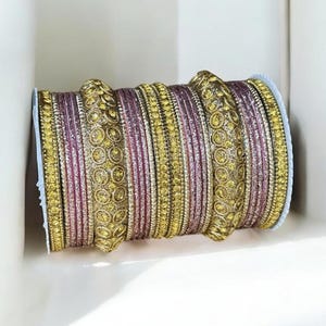 May include: A collection of ornate bangles in shades of gold and pink, stacked together. The bangles feature intricate patterns and embellishments, creating a luxurious and eye-catching design. The jewelry is displayed against a white background.