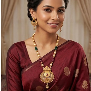 May include: A gold necklace featuring a large, ornate circular pendant with a central red stone and a small green drop, accented by beads in gold, green, and maroon. Matching gold drop earrings with a similar design are worn with the necklace.