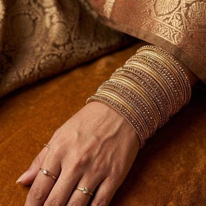 May include: A stack of gold-toned bangles with textured surfaces and embedded crystals worn on the wrist, accompanied by two thin gold rings featuring small clear stones on the fingers, showcasing a metallic finish and intricate detailing.