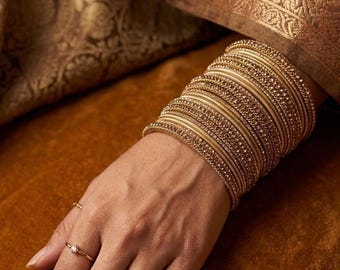 May include: A stack of gold-toned bangles with textured surfaces and embedded crystals worn on the wrist, accompanied by two thin gold rings featuring small clear stones on the fingers, showcasing a metallic finish and intricate detailing.