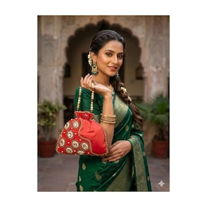 May include: A woman wearing a green sari and gold jewelry holds a red beaded purse with a pearl handle. The purse has gold and white embellishments. The woman is smiling and looking at the camera.