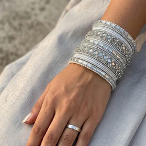 May include: A stack of silver-toned bangles featuring intricate geometric patterns, embedded with clear crystals and mirrored accents, worn alongside a simple silver band ring with a single small embedded stone on the finger.