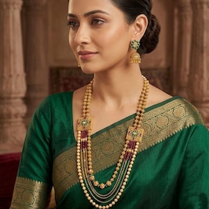 May include: A multi-strand gold necklace featuring two square pendants with green and red accents, connected by rows of gold beads and small red beads. Matching gold drop earrings with green and red circular centers complement the necklace.