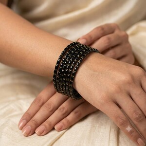 May include: A set of five black bangles featuring a faceted surface texture and small gold-colored metallic accents embedded along the edges. The bracelets are arranged in a stack, creating a uniform appearance with a dark, reflective finish.