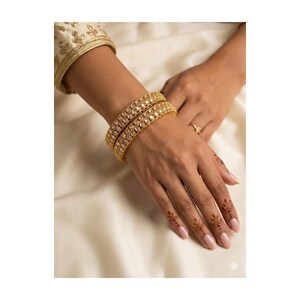 May include: Two gold bangles with clear stones are worn on a wrist, with a matching ring on a finger. The bangles are set against a cream-colored fabric. The sleeve of a gold embroidered garment is visible.