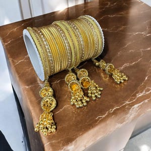 May include: A set of gold bangles with dangling charms. The bangles are decorated with small, sparkling stones. The charms are made of gold and have a bell shape.