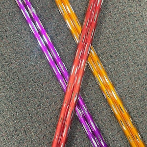 May include: Three colorful, metallic, and textured baton-like objects are arranged in an X-shape. The batons are red, purple, and orange, with silver accents. Small bells are attached to the ends. The objects are likely used for juggling or performance.
