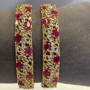 May include: Two gold bangles encrusted with alternating ruby-red and clear, oval-cut gemstones. The bangles are displayed on a gray surface, showcasing their intricate design and vibrant colors. The jewelry is designed to be worn as a set.