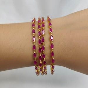May include: Four gold bangles, each adorned with oval-shaped, deep red gemstones. The gemstones are set in a line, connected by small gold beads. The bangles are displayed on a person's arm against a white background.