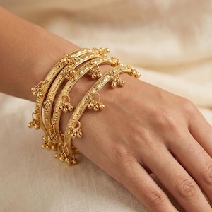 May include: A set of gold bangles adorns a wrist. Each bangle features intricate etched patterns and small dangling bell charms. The bangles are a warm gold color, reflecting light. The background is a soft, neutral tone.